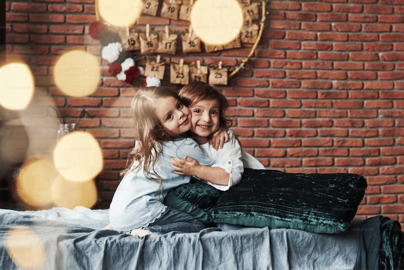 two children embracing on a bed with a brick wall behind them